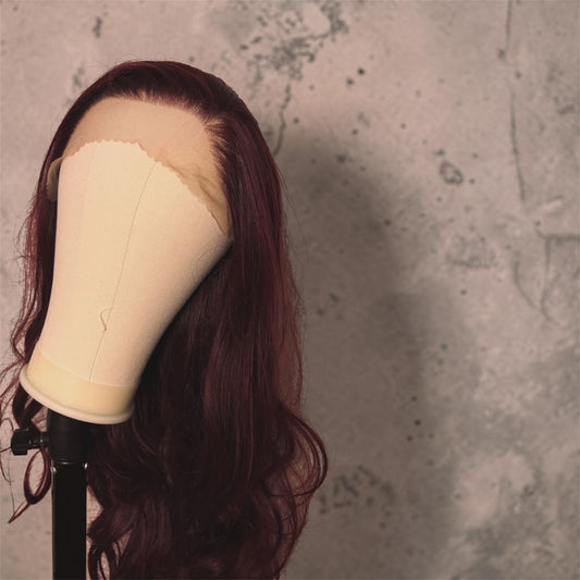A long wavy wig in deep burgundy with a beige lace front, displayed on a mannequin head against a textured gray concrete wall.