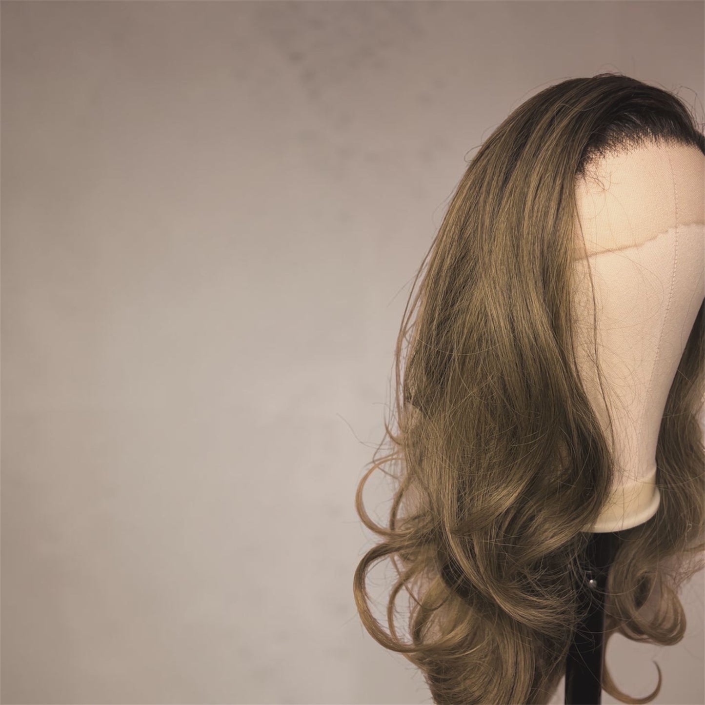 A wavy, shoulder-length wig with warm brown and blonde highlights, displayed on a mannequin head against a neutral beige background.